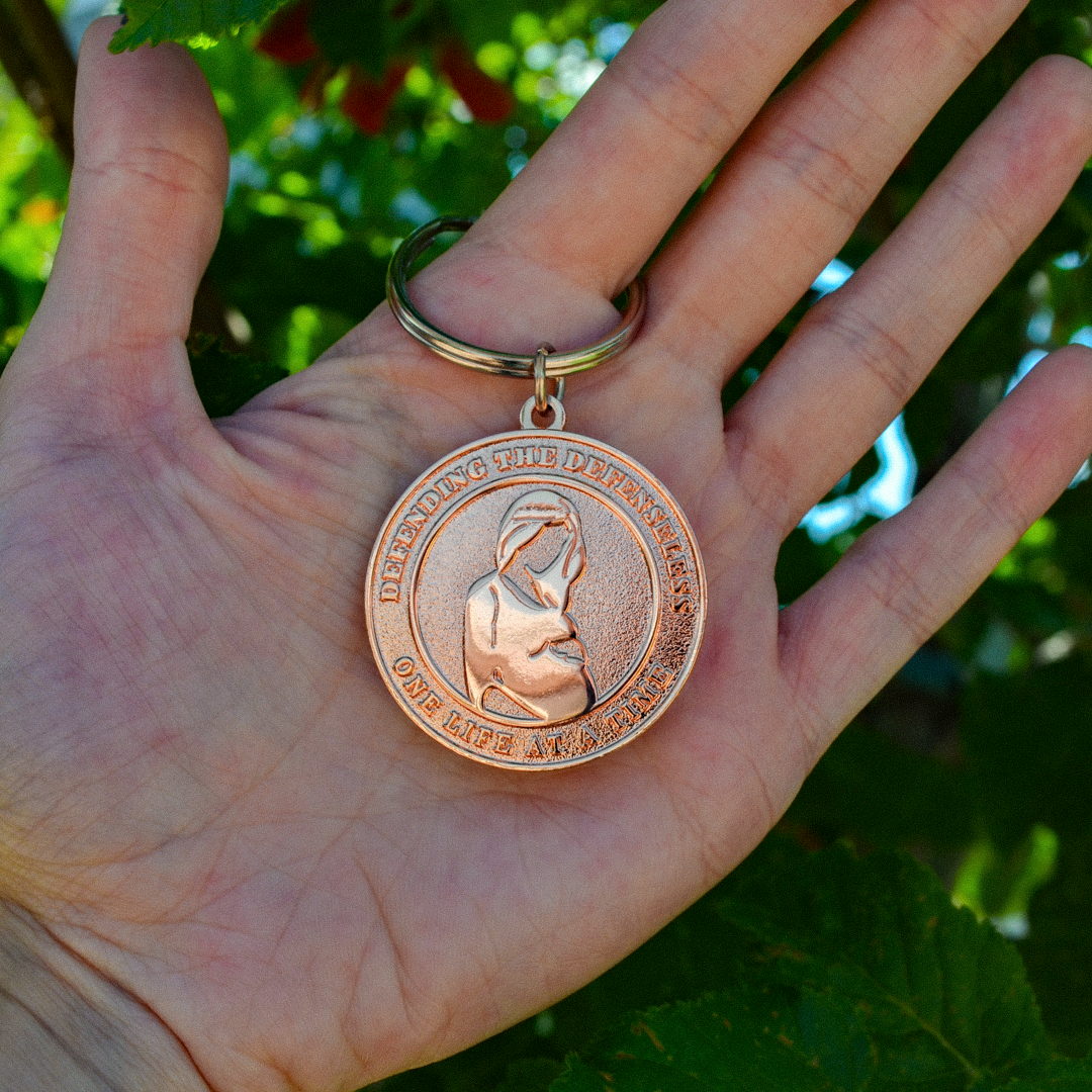 A round, rose gold-colored 1000 Miracles commemorative keychain medallion held in a person's hand, featuring an embossed pregnant woman and the text 'DEFENDING THE DEFENSELESS ONE LIFE AT A TIME' on a key ring.