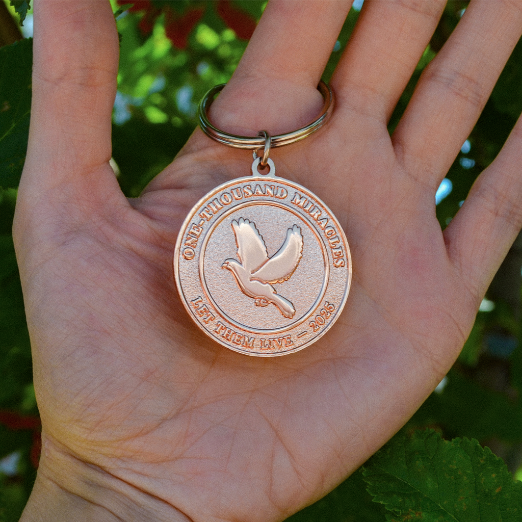 A round, rose gold-colored 1000 Miracles commemorative keychain medallion held in a person's hand, featuring an embossed dove and the text 'ONE THOUSAND MIRACLES, LET THEM LIVE - 2025' on a key ring.