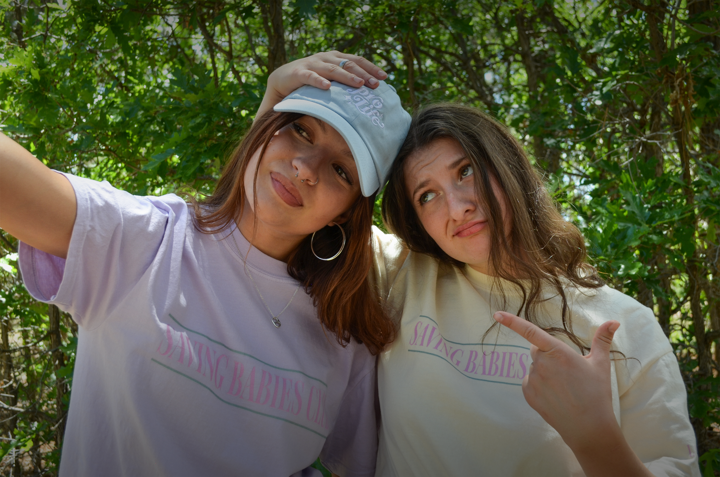 Two women wearing matching pro-life t-shirts with "Saving Babies Club" text in a forest setting