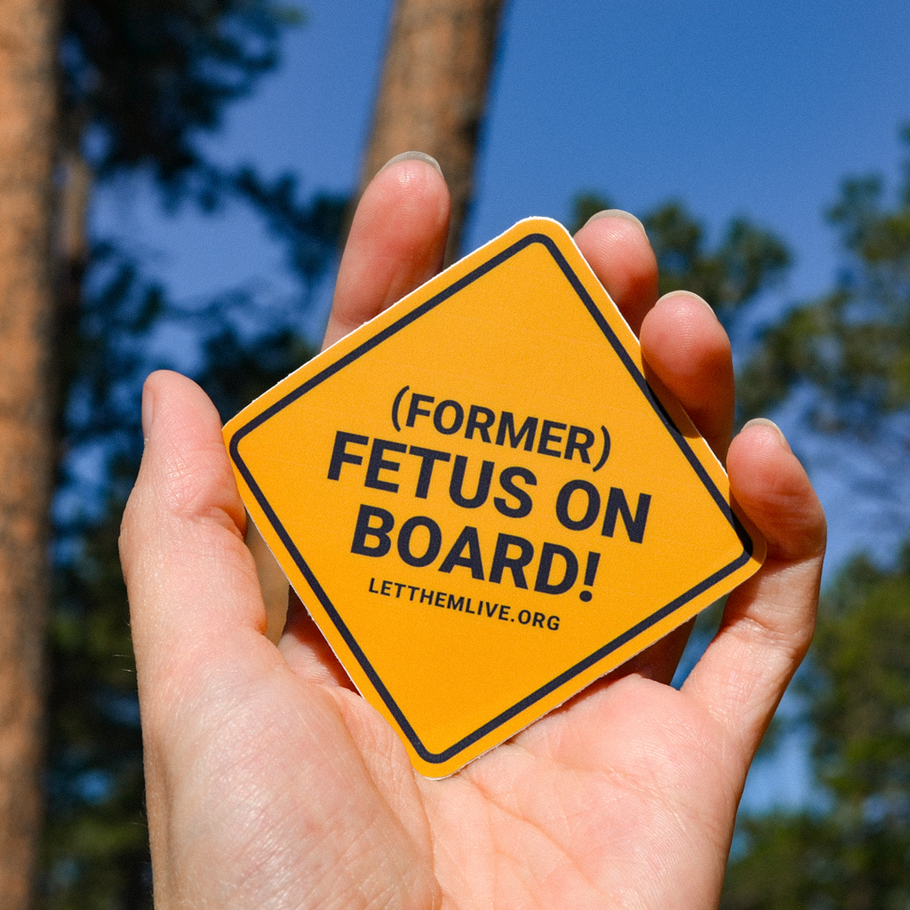 Hand holding orange pro-life sticker with the text "Former Fetus On Board"