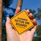 Hand holding orange pro-life sticker with the text "Former Fetus On Board"