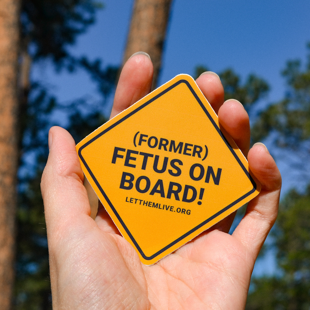 Hand holding orange pro-life sticker with the text "Former Fetus On Board"
