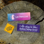 Assortment of three pro-life stickers on tree stump, including "Former Fetus On Board", "Let Them Live Logo", and "It's Okay to Like Babies Being Born"