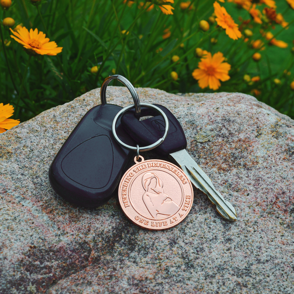 A round, rose gold-colored 1000 Miracles commemorative keychain medallion on a rock, featuring an embossed pregnant woman and the text 'DEFENDING THE DEFENSELESS ONE LIFE AT A TIME' on a key ring with car keys.
