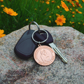 A round, rose gold-colored 1000 Miracles commemorative keychain medallion on a rock, featuring an embossed pregnant woman and the text 'DEFENDING THE DEFENSELESS ONE LIFE AT A TIME' on a key ring with car keys.