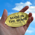Hand holding a gold oval with 'MADE IN THE WOMB' text against a blue sky.