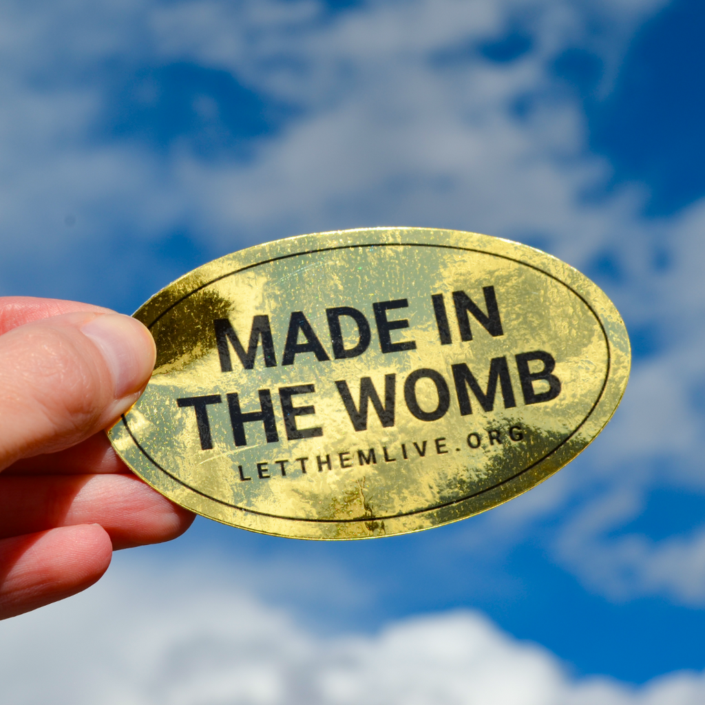 Gold oval sticker with 'MADE IN THE WOMB' text held against a blue sky.