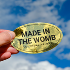 Gold oval sticker with 'MADE IN THE WOMB' text held against a blue sky.