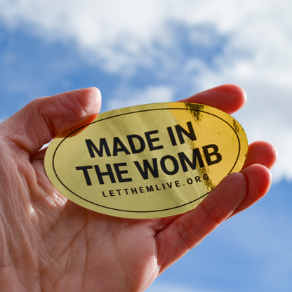 Hand holding a gold sticker with 'MADE IN THE WOMB' text against a blue sky.